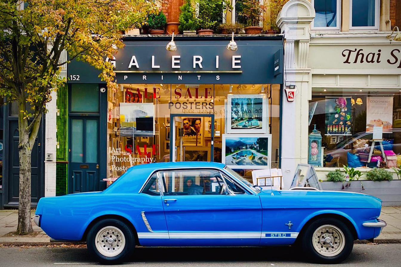 A classic blue Mustang parked outside the real Galerie Prints storefront at 152, autumn tree overhead, prints visible in the window